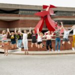 in action shot of students jumping and holding hands in front of sculpture