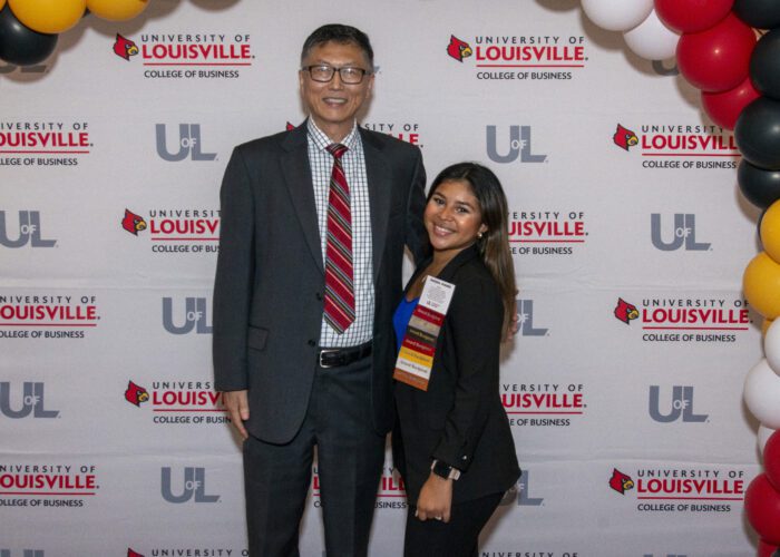 Justyne Aranda and Interim Dean Dr. Jeff Guan pose for a photo with the College of Business logo background and red, yellow, black and white balloons