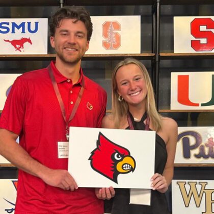 ACC InVenture 2024 competitor Eric Nelson poses with a teammate holding a sign with the UofL Cardinal head. Other university logos are on shelves behind them.