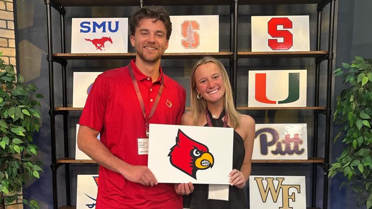ACC InVenture 2024 competitor Eric Nelson poses with a teammate holding a sign with the UofL Cardinal head. Other university logos are on shelves behind them.