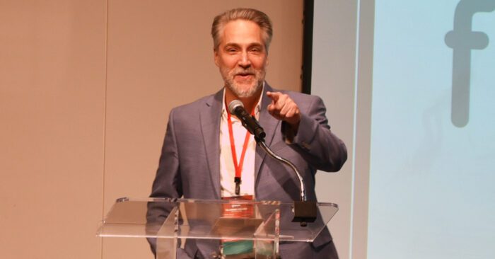 Scott Koloms, CEO of Facilities Management Services and founder of Canopy, speaks on stage in front of a projector screen at the Speed Art Museum as part of the 2024 Cardinal Challenge.