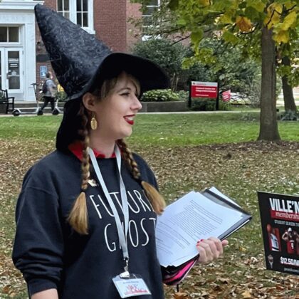 Katie Jo Glesing stands on a pathway on the Belknap campus, conducting a Creepy Campus Tour. She is wearing a black witch's hat and is holding a clipboard.