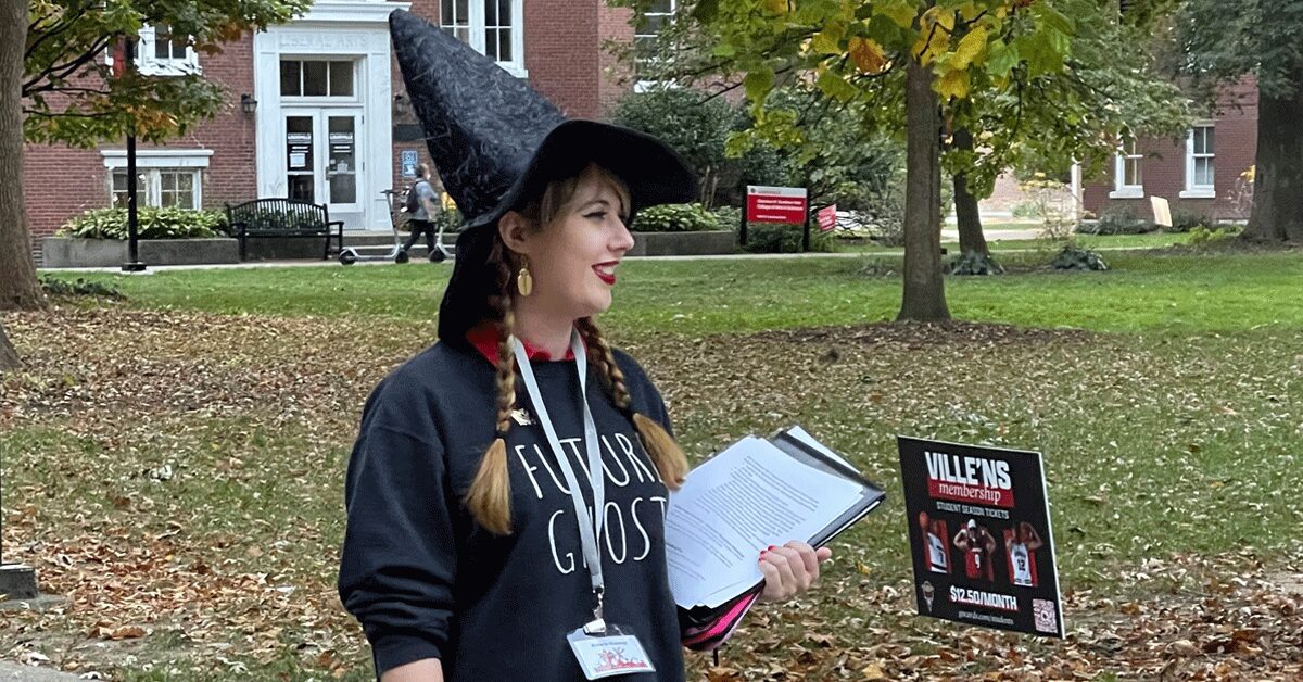 Katie Jo Glesing stands on a pathway on the Belknap campus, conducting a Creepy Campus Tour. She is wearing a black witch's hat and is holding a clipboard.