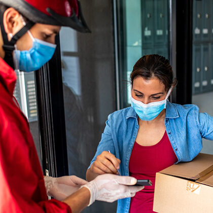Рабочий в красной куртке, велосипедном шлеме, защитной маске и хирургических перчатках во время пандемии Covid-19 доставляет посылку женщине в маске.