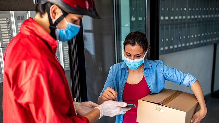 Ein wichtiger Arbeiter, der während der Covid-19-Pandemie eine rote Jacke, einen Fahrradhelm, eine schützende Gesichtsmaske und OP-Handschuhe trägt, liefert ein Paket an eine maskierte Frau.