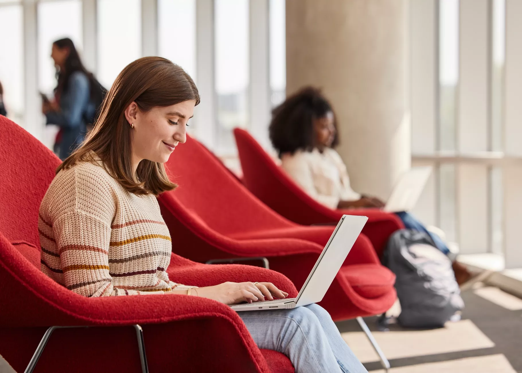Lexi Raikes sitting on the second floor of the Belknap Academic Building, in a red chair, typing happily on her laptop