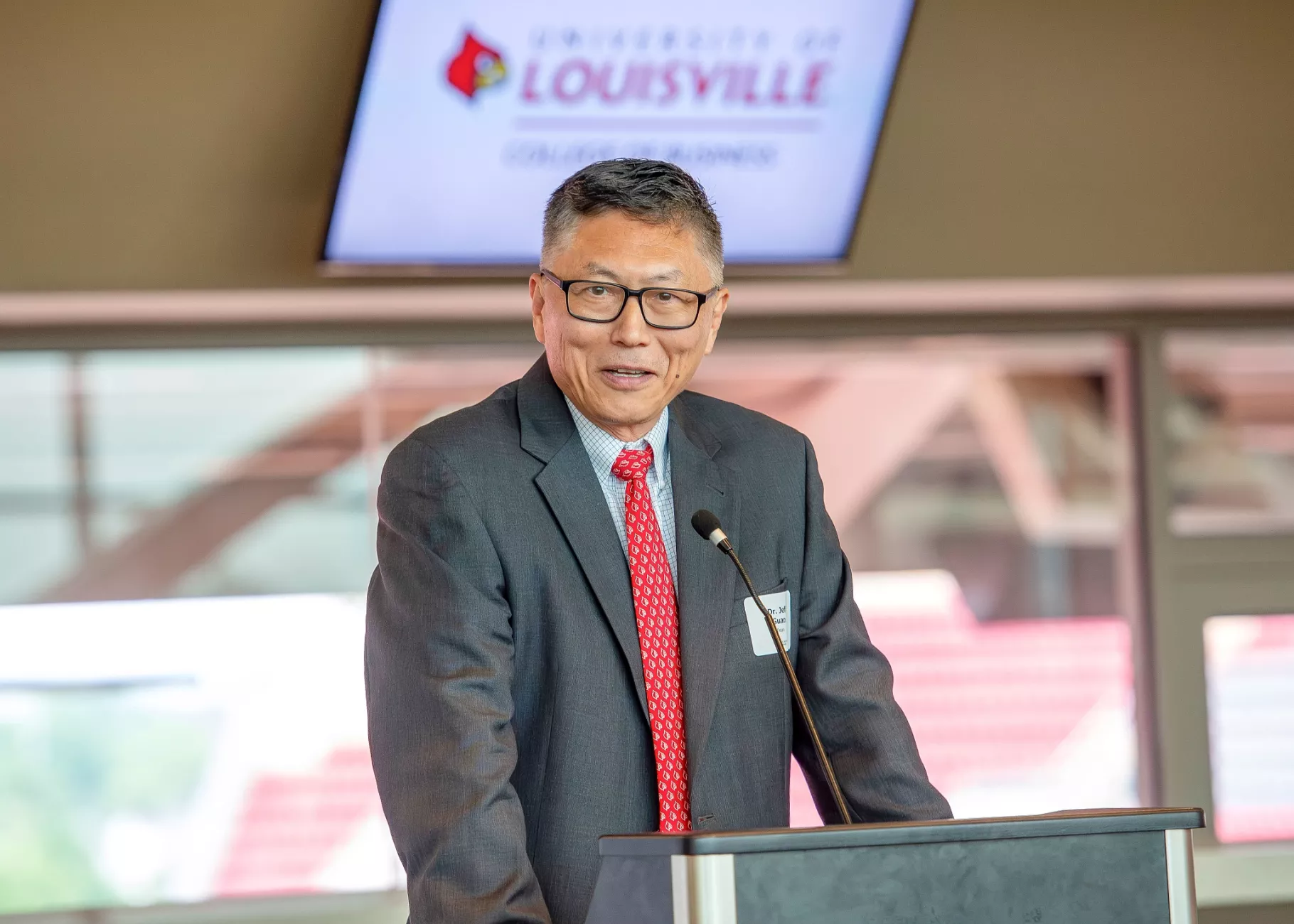 UofL College of Business Interim Dean, Jeff Guan, gives an opening speech at a podium of the PNC club with a CoB logo on a screen behind him.