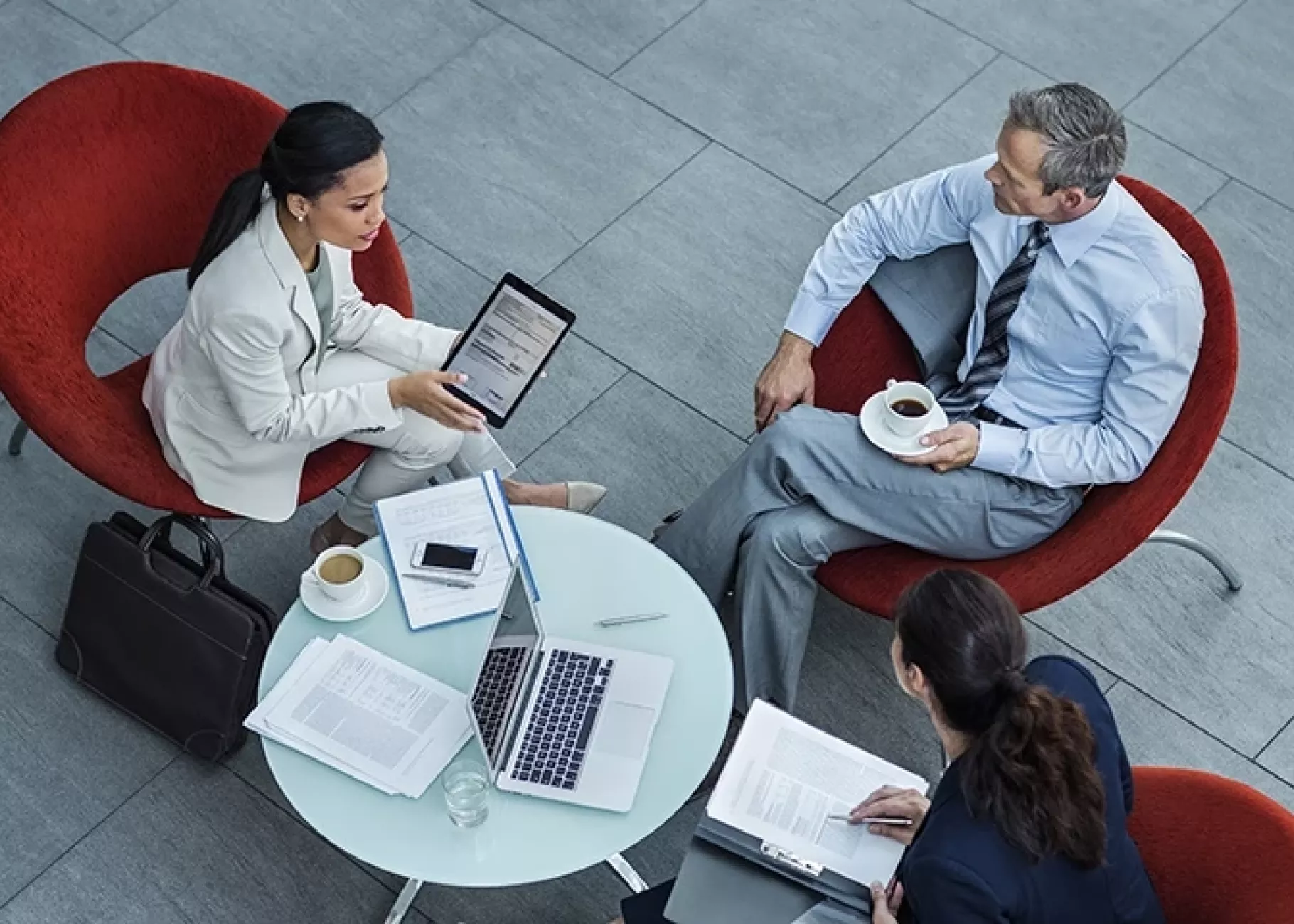 Three people in business attire sitting around a table with coffee, laptops, tablets, and notebooks, engaged in conversation.