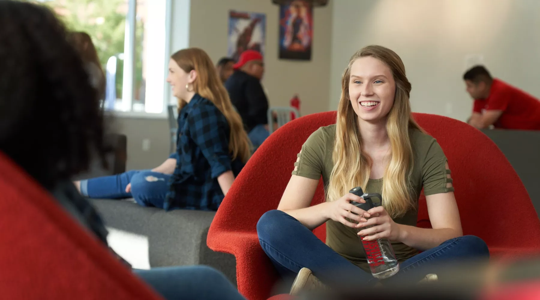 Student sitting in a red chair and talking with another student while holding a water bottle.