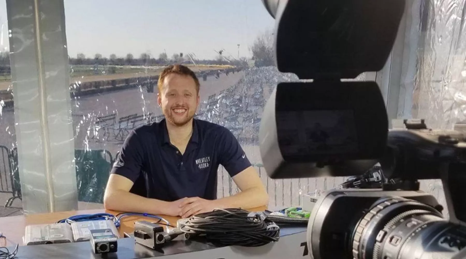 College of Business Equine student prepares for a broadcast in front of a camera with a horse racing track in the background.