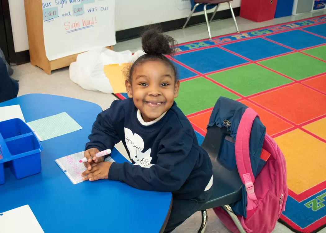 A young girl with a ponytail missing her two front teeth smiling big at the camera while sitting at her desk writing with a pink marker