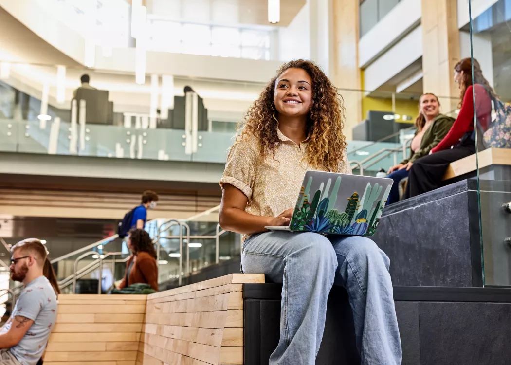 Student recognizes another student walk into the Belknap Academic Building as she is typing on her laptop and sitting on the wooden platform benches at the entrance of the BAB.