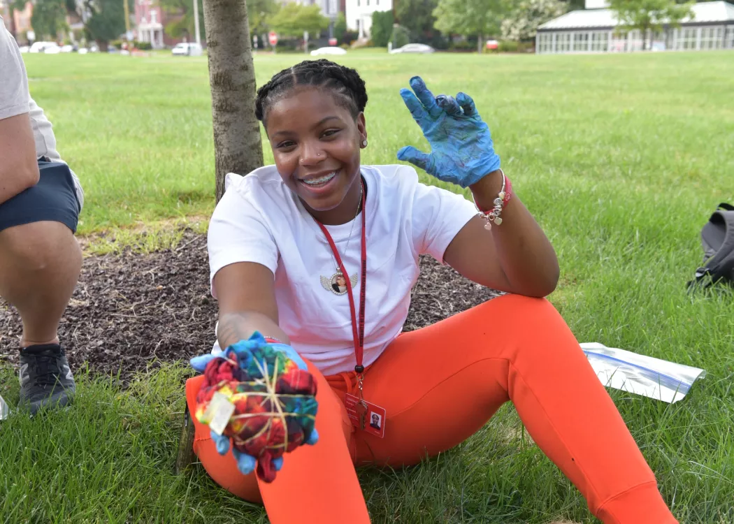 TILE student holding out tie-dyed shirt and holding up "L" hand symbol while sitting in grass on University of Louisville campus in front of the College of Business.