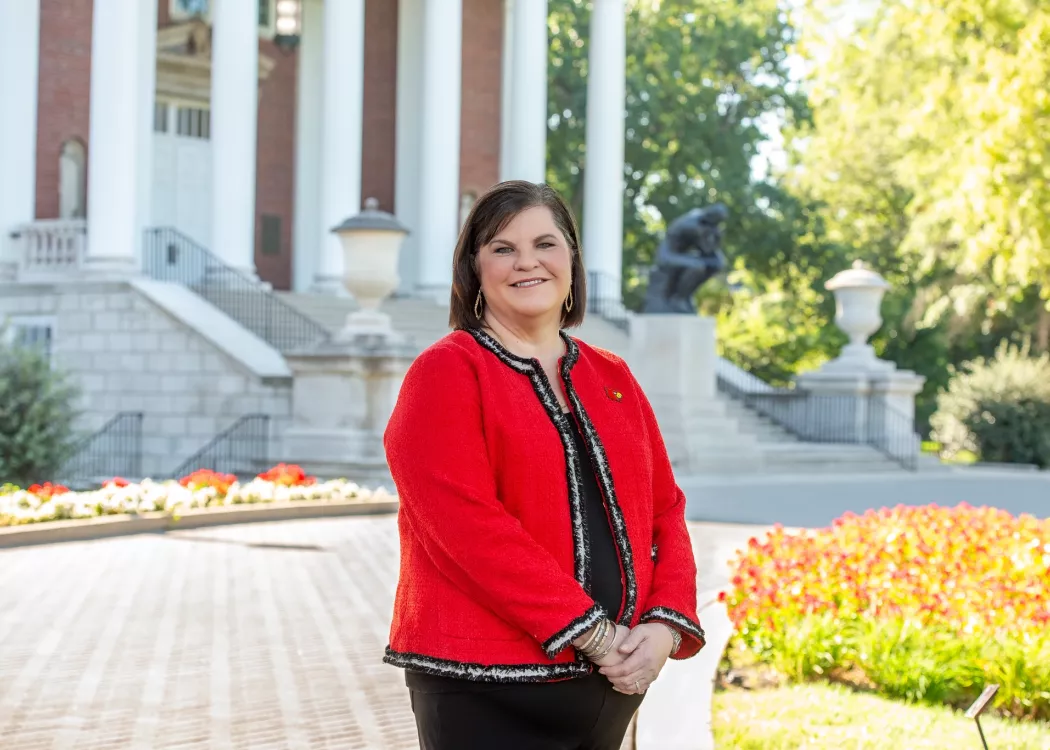 Woman in Cardinal red blazer standing in front of building with columns and bronze statue of The Thinker on UofL's Belknap…