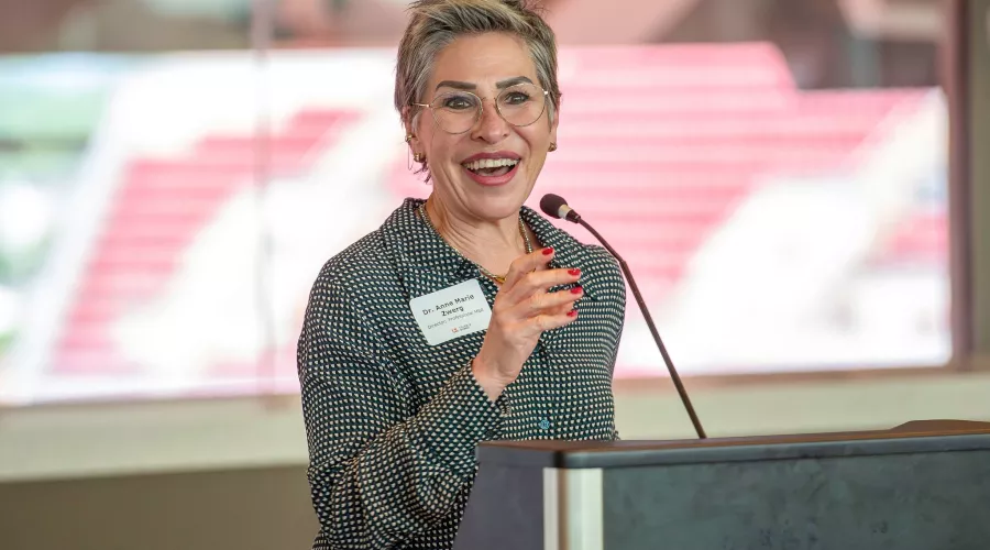 UofL College of Business MBA Program director, Anne Zwerg,  gives an opening speech at a podium of the PNC club. A window is in the background that look out onto the stands of Cardinal Stadium.