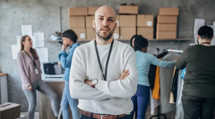 Project Manager crosses arms in the foreground, looking at the camera, while employees sort through merchandise in the background.