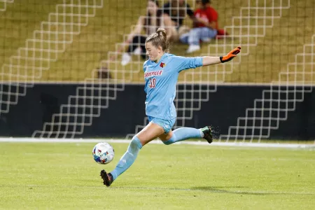 Female goalie in blue UofL uniform posed to kick soccer ball